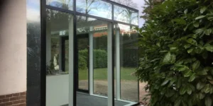 Serre als uitbouw met zwart stalen kozijnen en glazen pui aan een woning, met zicht op de tuin