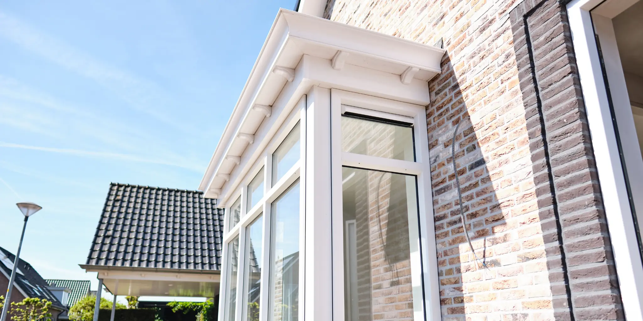 Witte erker met afgeschuind dak en sierlijke daklijst tegen een bakstenen gevel, gefotografeerd van onderaf tegen een blauwe lucht