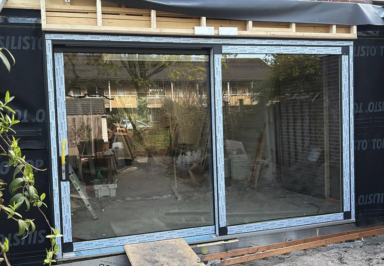 Nieuwe kozijnen met schuifpui geplaatst in een aanbouw in aanbouwfase, met isolatiefolie en bouwmateriaal nog zichtbaar rondom het glas