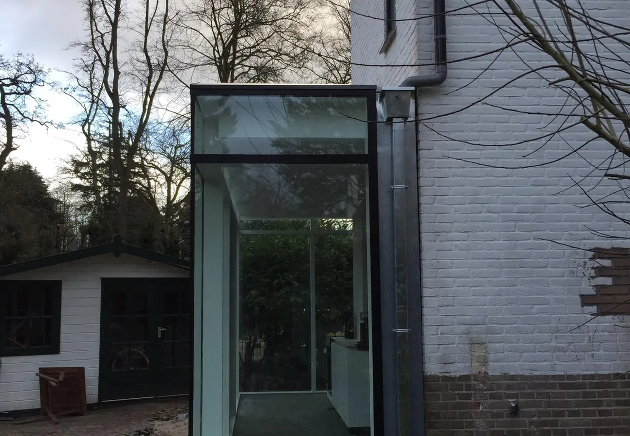 Aanbouw met glazen hoekconstructie tegen een wit geschilderde gevel, met een houten schuur en kale bomen op de achtergrond
