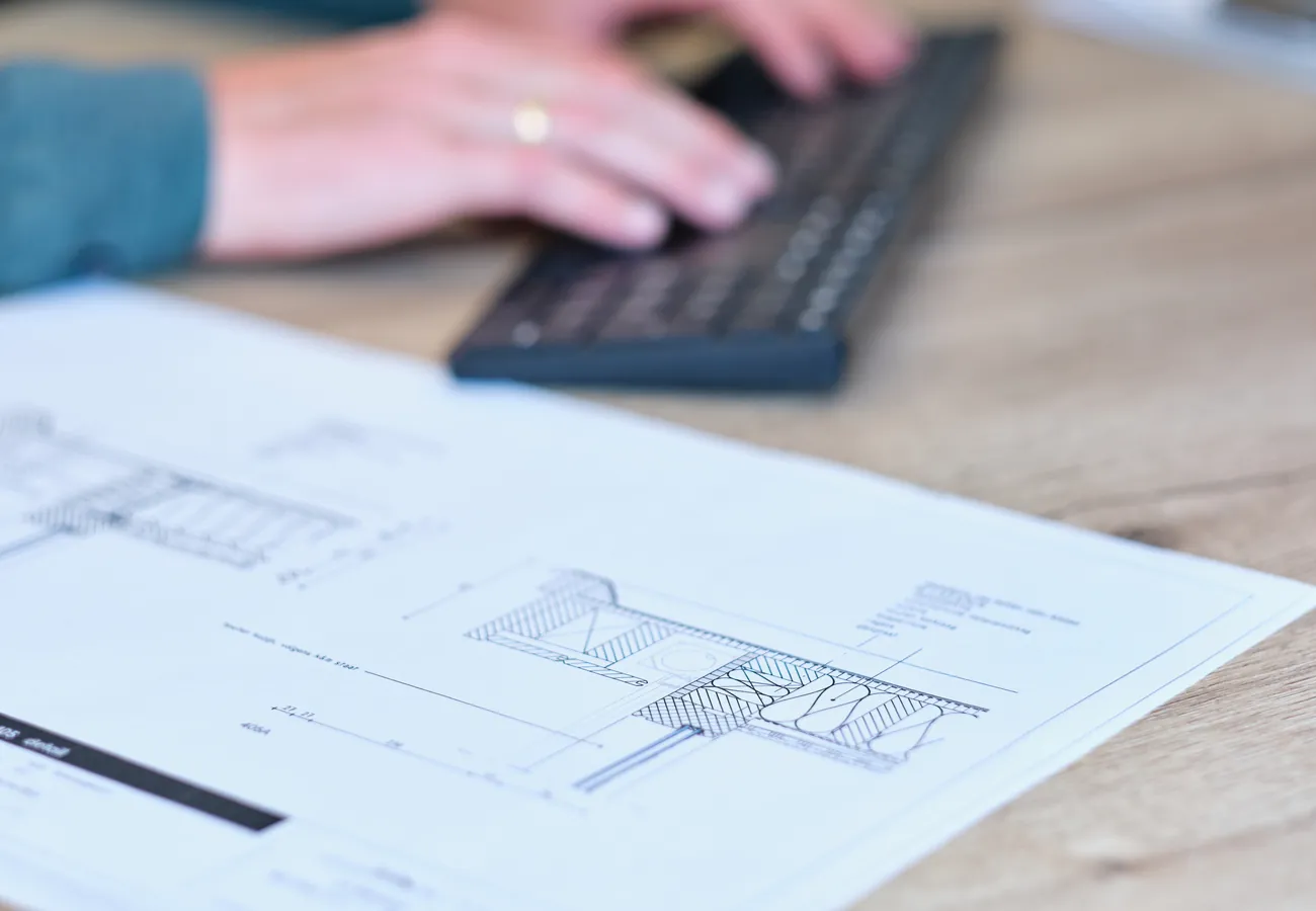 Detailtekening van een aanbouw op tafel, terwijl iemand de offerte of planning uitwerkt op de computer
