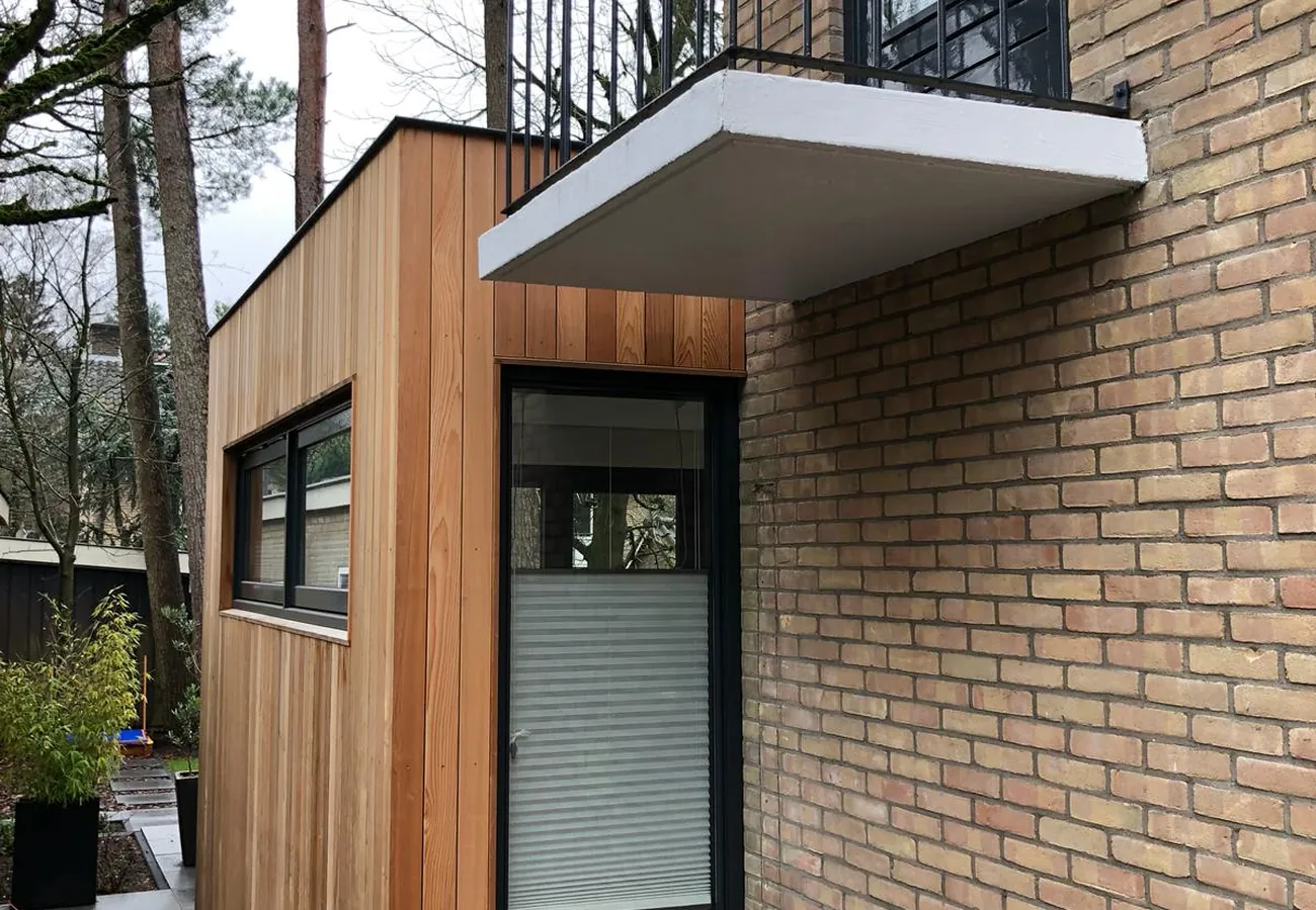 Houten uitbouw tegen de zijgevel van een woning, met een balkon op de verdieping erboven