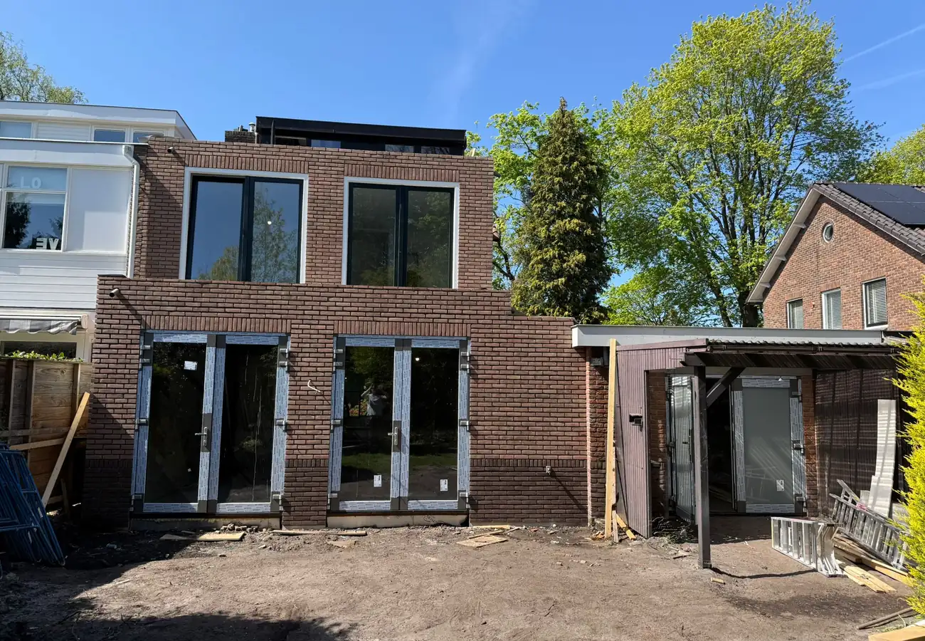 Afgebouwde aanbouw als thuiskantoor met bakstenen gevel, grote raampartijen en openslaande tuindeuren, gefotografeerd op een zonnige dag in de afrondende bouwfase.