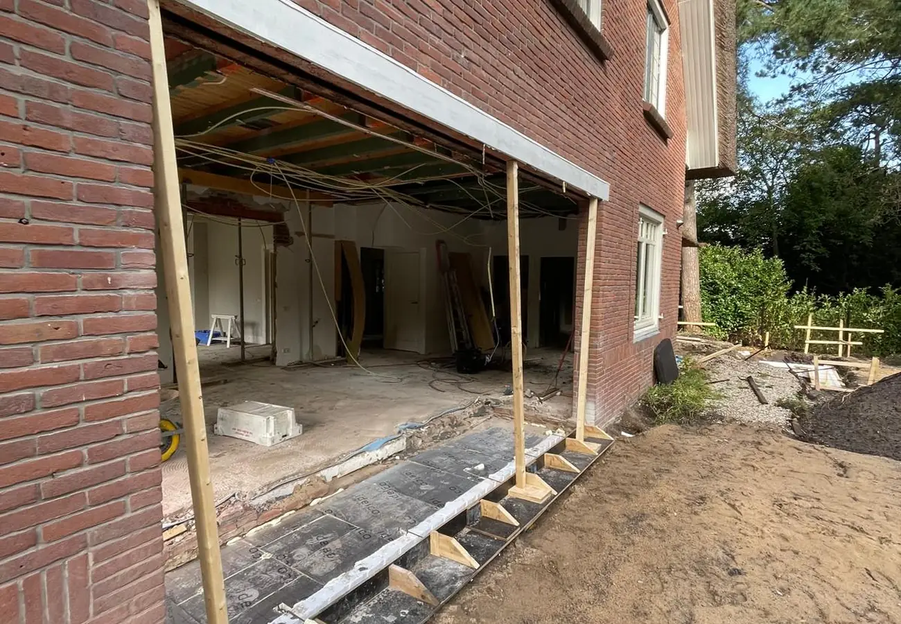 Close-up van een opengebroken gevel bij de bouw van een aanbouw, met tijdelijke steunbalken, isolatieplaten op de fundering en zicht op de bekabeling in de vloerconstructie.